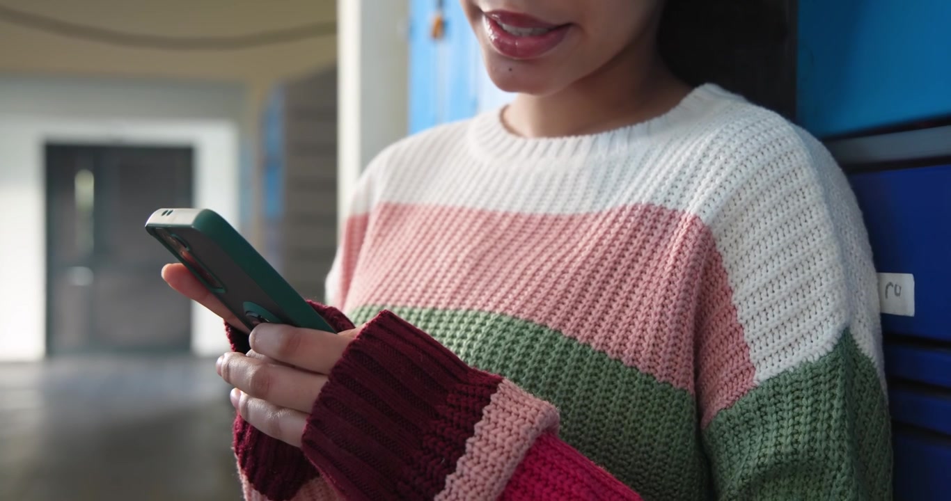 Alunna che indossa un maglione di lana, appoggiata ad armadietti blu, che manda messaggi agli amici con un telefono verde