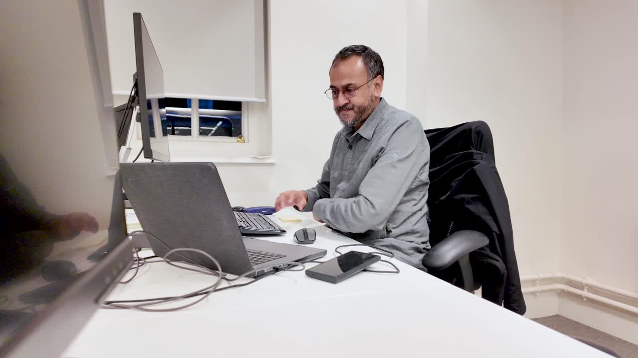 Hombre del sur de Asia poniéndose las gafas en un escritorio de una oficina moderna con un portátil y un monitor de ordenador. Concepto de negocio, teletrabajo, concentración y enfoque.