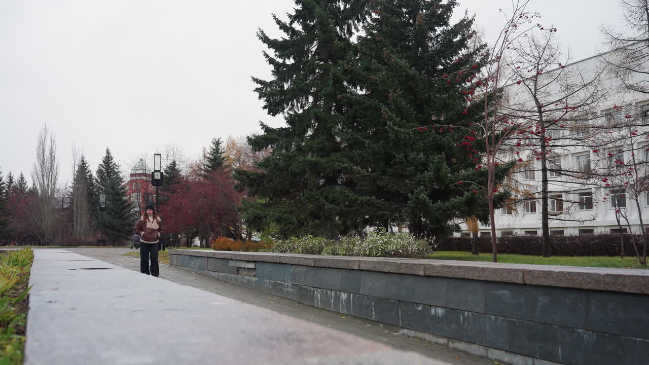 Thoughtful girl wearing black cap brown jacket and trouser walking slowly with hands in pockets on a pathway surrounded by pine trees flower beds modern buildings and overcast sky during cold autumn