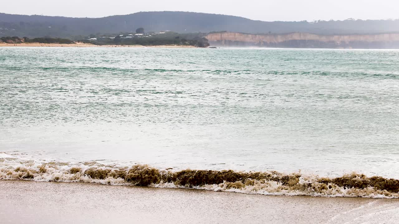 Calm ocean waves roll onto the sandy beach under overcast skies at Great Ocean Road, Australia, creating a serene coastal scene