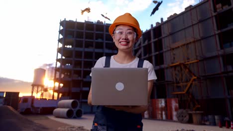 trabajadora asiática con gafas de protección y casco de seguridad usando una computadora portátil y apuntando hacia un lado mientras está de pie en el sitio de construcción