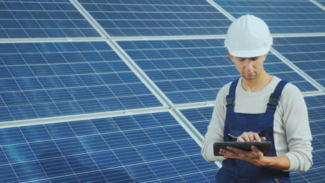 young engineer in white helmet works with a tablet on the background of solar panels