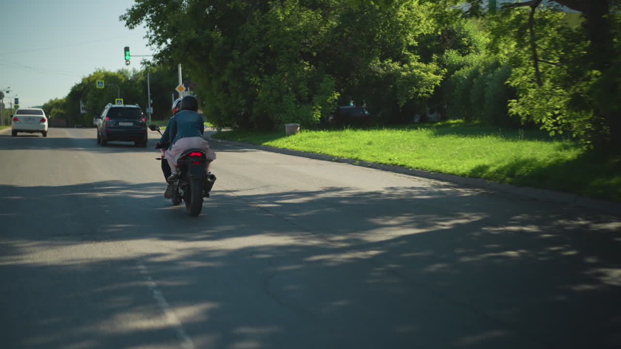 전동 자전거를 타는 두 친구의 뒷면, 앞에 초록색 신호등 지표가 있는 두 차를 추적하고, 빛의 날에 그늘진 도시 도로를 따라 여행하는 동안 트램도 에서 볼 수 있습니다.