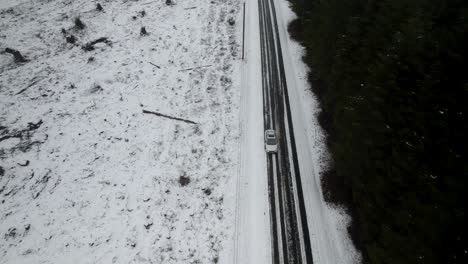 Ascending-up-over-a-white-car-traveling-along-a-snow-covered-lonely-country-road,-aerial