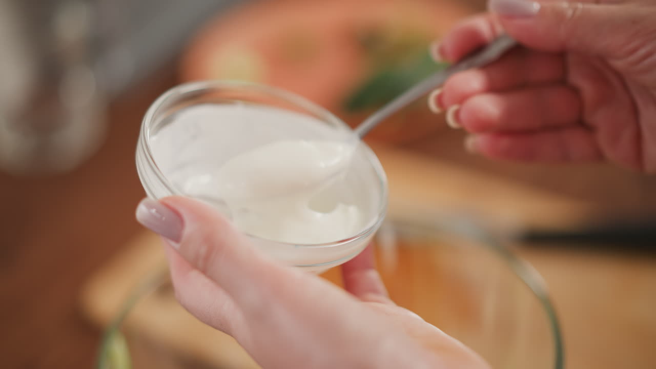 close up of person holding transparent glass bowl filled with creamy salad dressing while gently scooping with metallic spoon in well-lit kitchen with wooden countertop and soft blur in background