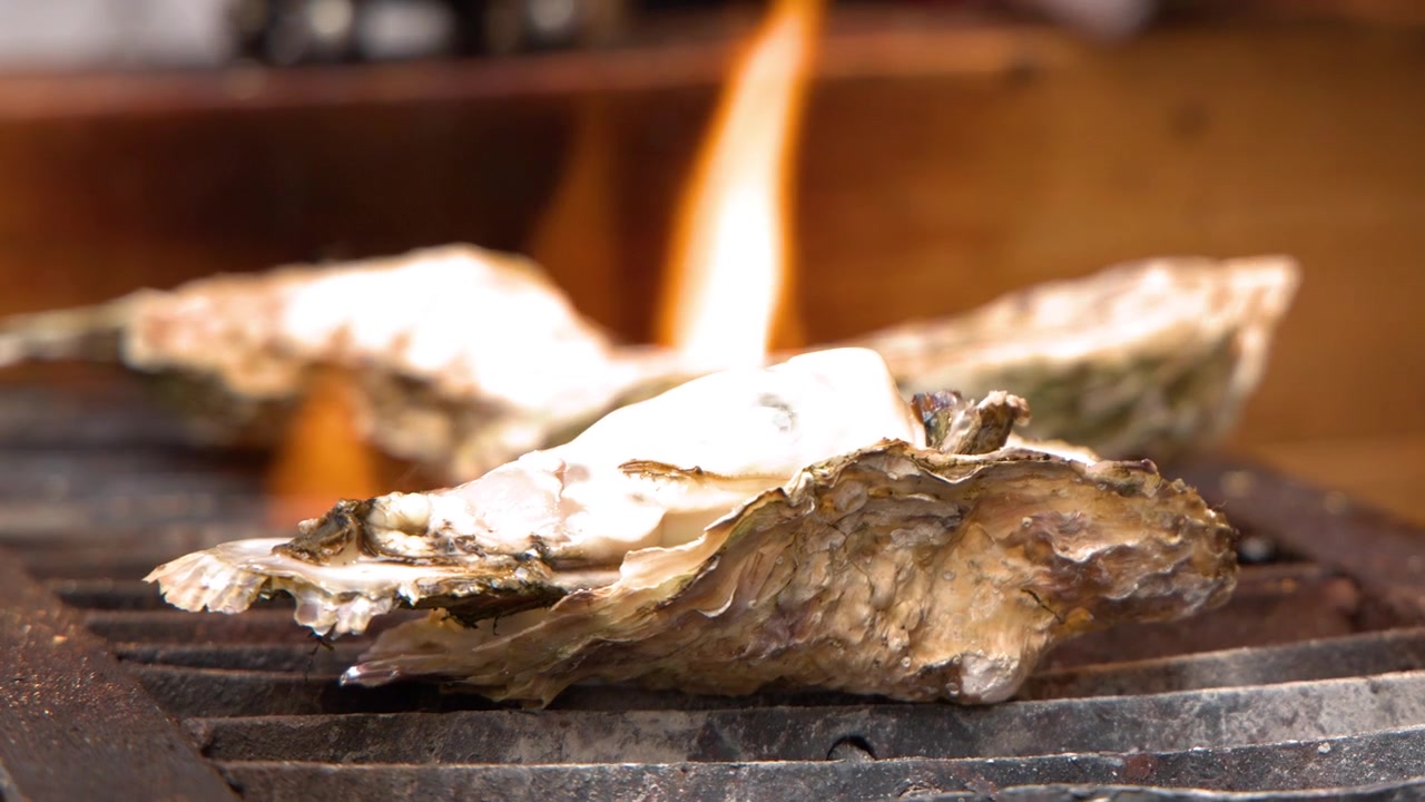 Close-up shot of large oysters cooking on a grill with flickering orange flames in Tokyo.