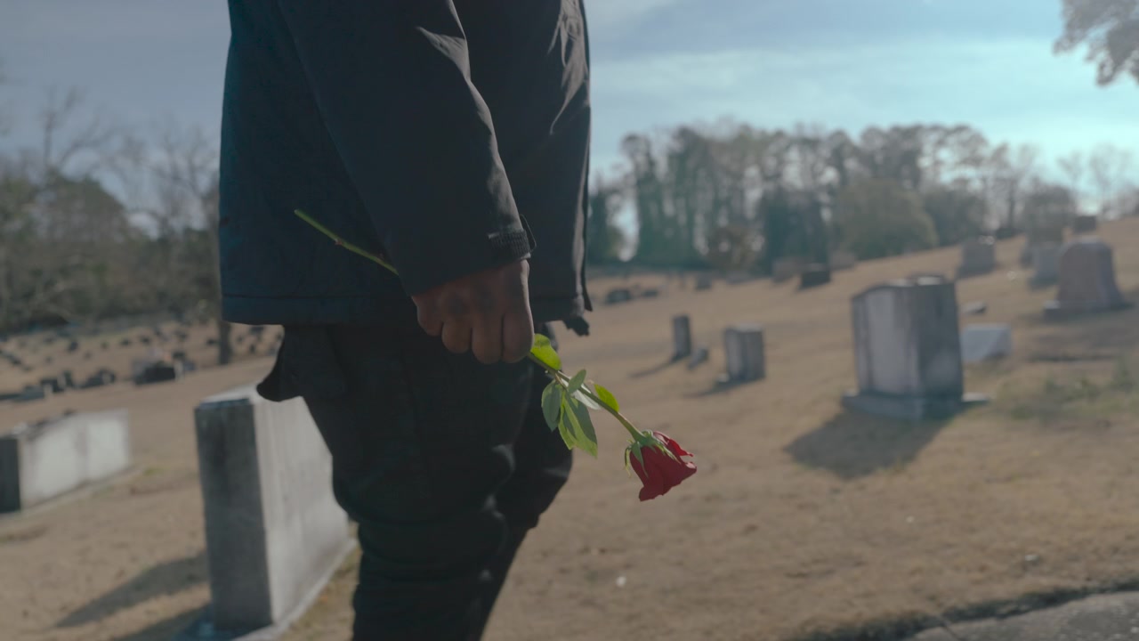 Uomo in lutto che tiene in mano una singola rosa rossa mentre cammina tra le lapidi in un cimitero