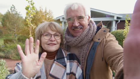 Fröhlicher-älterer-Mann-Und-Frau-In-Stilvoller,-Warmer-Freizeitkleidung,-Die-Sie-Mit-Einem-Lächeln-Betrachten,-Während-Sie-Die-Kamera-Vor-Garten-Und-Haus-Begrüßen