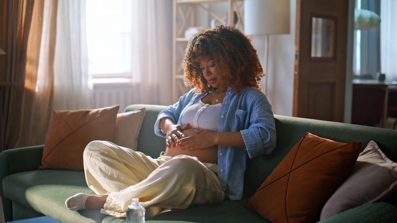 Futura mamma che applica olio sulla pancia incinta sul divano. Mamma che si prende cura della pancia in soggiorno
