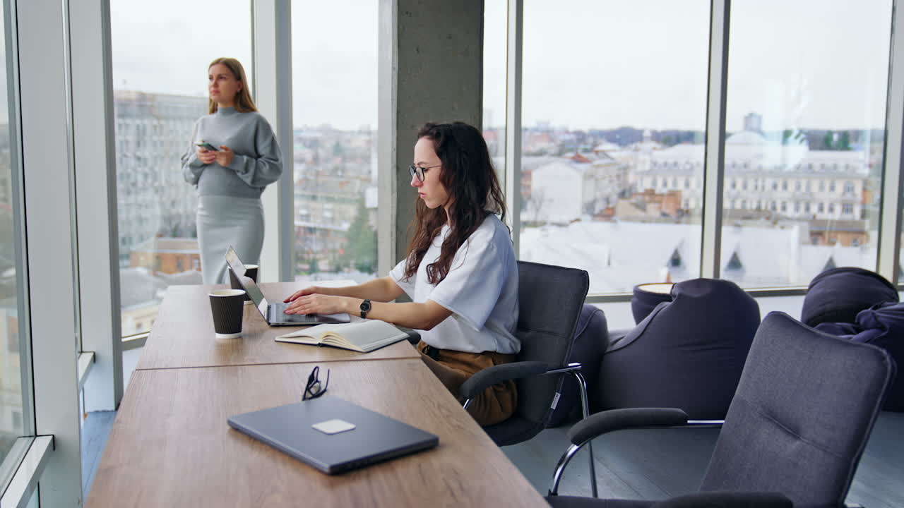 Una donna bruna seria si mette gli occhiali e inizia a lavorare al suo laptop. Una collega incinta sullo sfondo è concentrata sul suo telefono