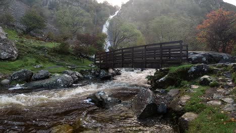 Cascadas-De-Agua-Que-Fluyen-Rápidamente-Y-Poderosas-A-Cámara-Lenta-Bajo-Un-Puente-De-Madera-Desde-El-Desierto-De-La-Cascada-Del-Valle-Del-Bosque