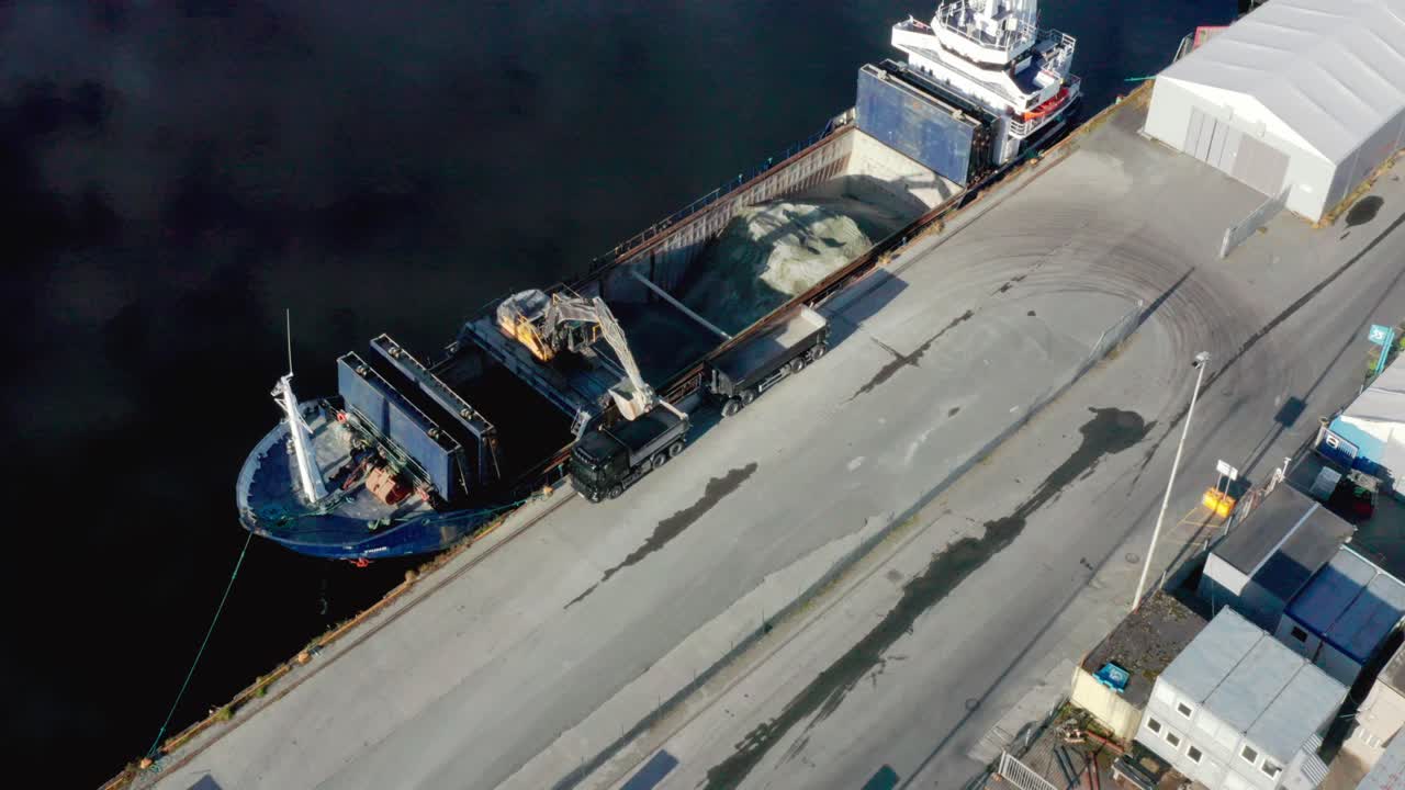 High-angle top-down aerial orbit shot of an excavator unloading gravel from a barge onto trucks at an industrial harbor. Provides a clear overview of construction logistics, heavy machinery workflow,