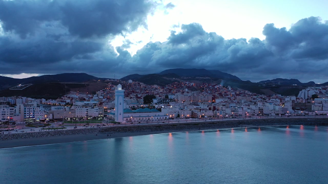 Ampia vista aerea all'ora blu di M'diq sulla costa mediterranea del Marocco, con una moschea sul lungomare, mare calmo, luci della città, i Monti del Rif e nuvole serali spettacolari.