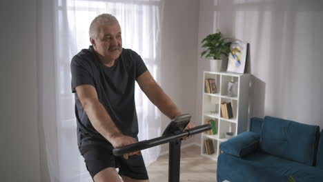 tired elderly man is spinning pedals of stationary bicycle training alone in home keeping physical activity healthy lifestyle