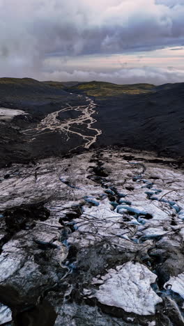 Flight above the old glacier descending among the black rocks. Dramatic cloudscape covering the sky at backdrop. Iceland scenery. Vertical video.
