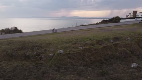 Cars-overtaking-cyclist-riding-on-hilly-road-upwards-during-cloudy-day-in-Uruguay---Aerial-tracking-shot