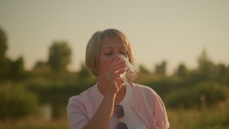 mujer de mediana edad con camisa rosa disfrutando de una refrescante copa de agua al aire libre bajo un cielo soleado, ella sostiene el vaso con una expresión relajada, saboreando el momento en un entorno verde natural