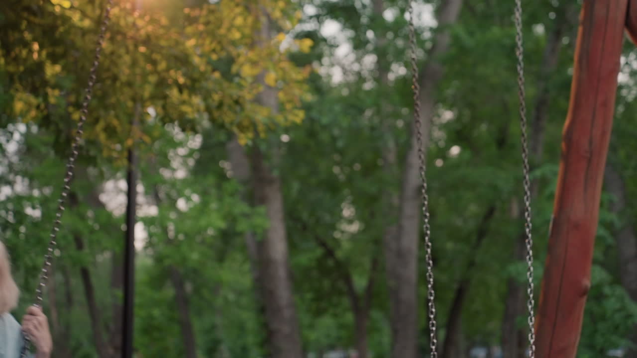 playful mom on swing in sunlit playground lifts hair as swing rocks back and forth laughter filling calm park dusk scene capturing joyful carefree moment of adult childlike fun amid green trees