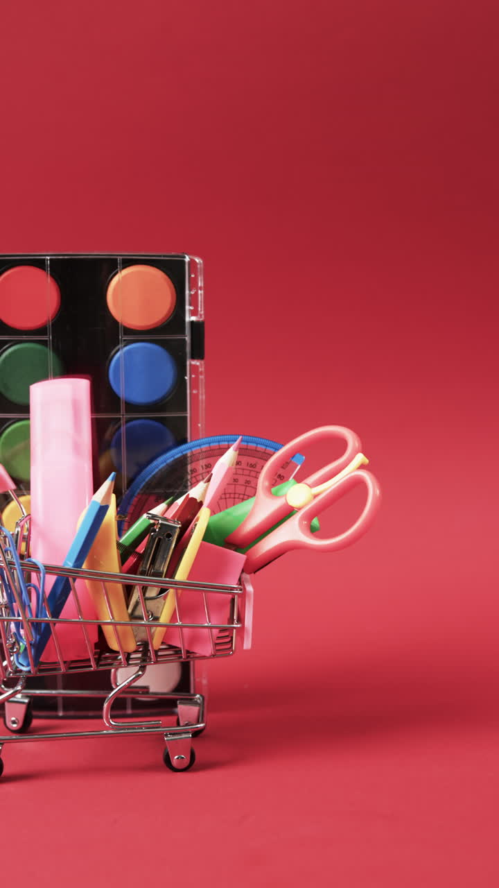 Vertical video of close up of shopping trolley with school items and paints on red background