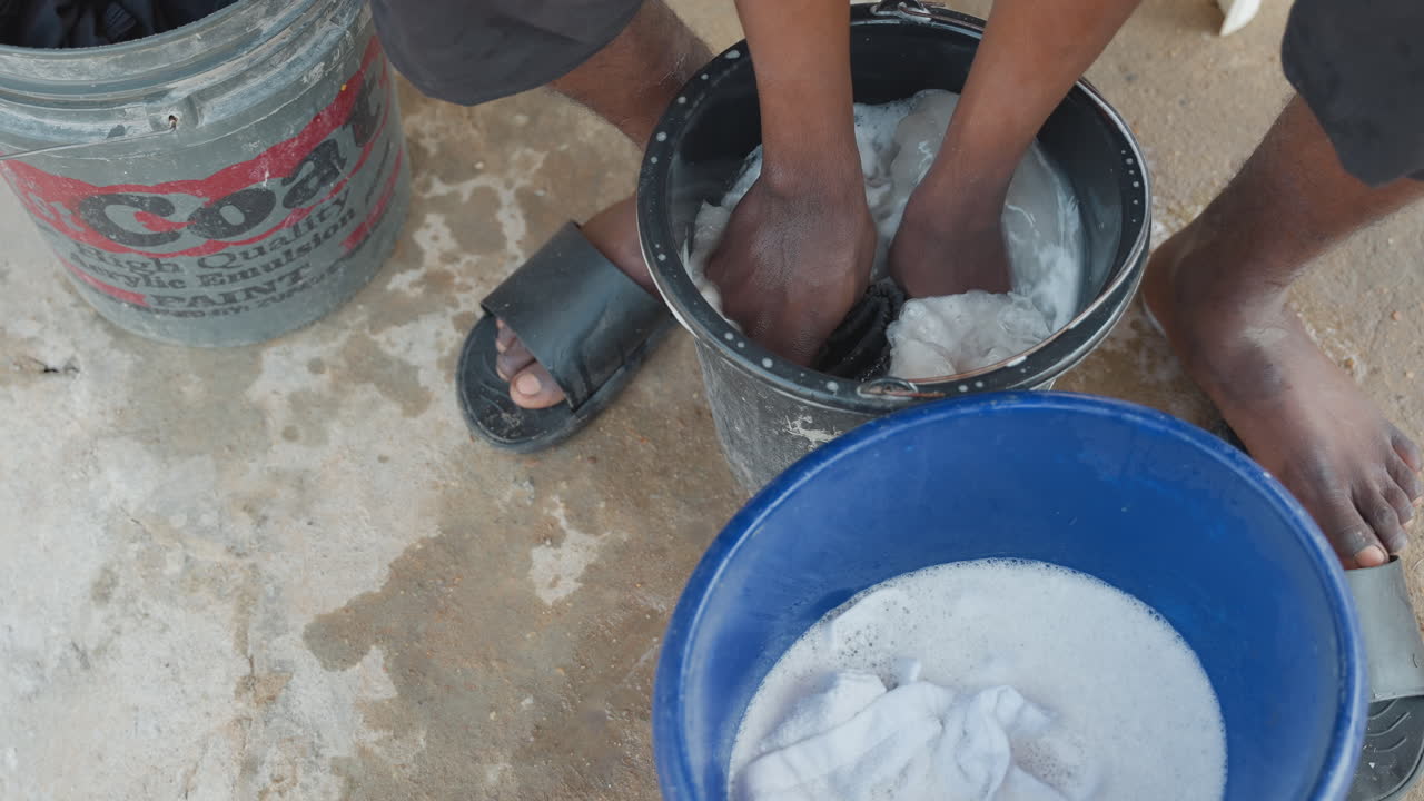 Mani nere lavano un capo scuro in un secchio blu. Una persona inginocchiata su un portico di cemento sfrega il tessuto con i palmi. Polvere e schiuma si mescolano, un movimento manuale concentrato cattura una faccenda domestica di routine.