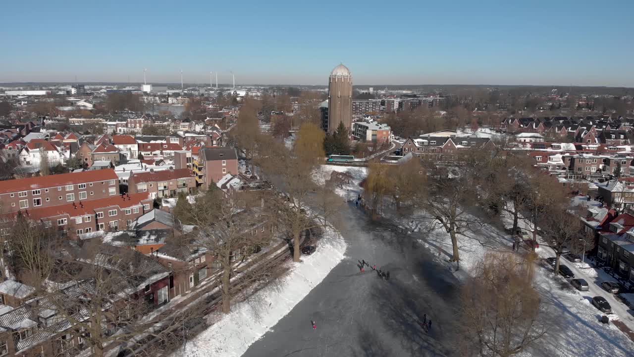 escena de invierno urbano aéreo lateral con gente patinando sobre hielo a lo largo del canal congelado curvo que atraviesa la ciudad holandesa de zutphen con sombras de árboles estériles y una antigua torre de agua en el fondo