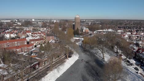 sideways aerial urban winter scene with people ice skating along the curved frozen canal going through the dutch city of zutphen with shadows of barren trees and former water tower in the background