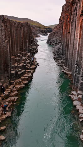 flying above the river flowing among the columnar rocks. tourists walk by the multiple boulders at the waterfront. iceland mountains. vertical video.