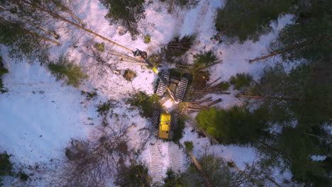 Overhead-drone-view-of-yellow-harvester-felling-trees-at-night-in-snowy-forest