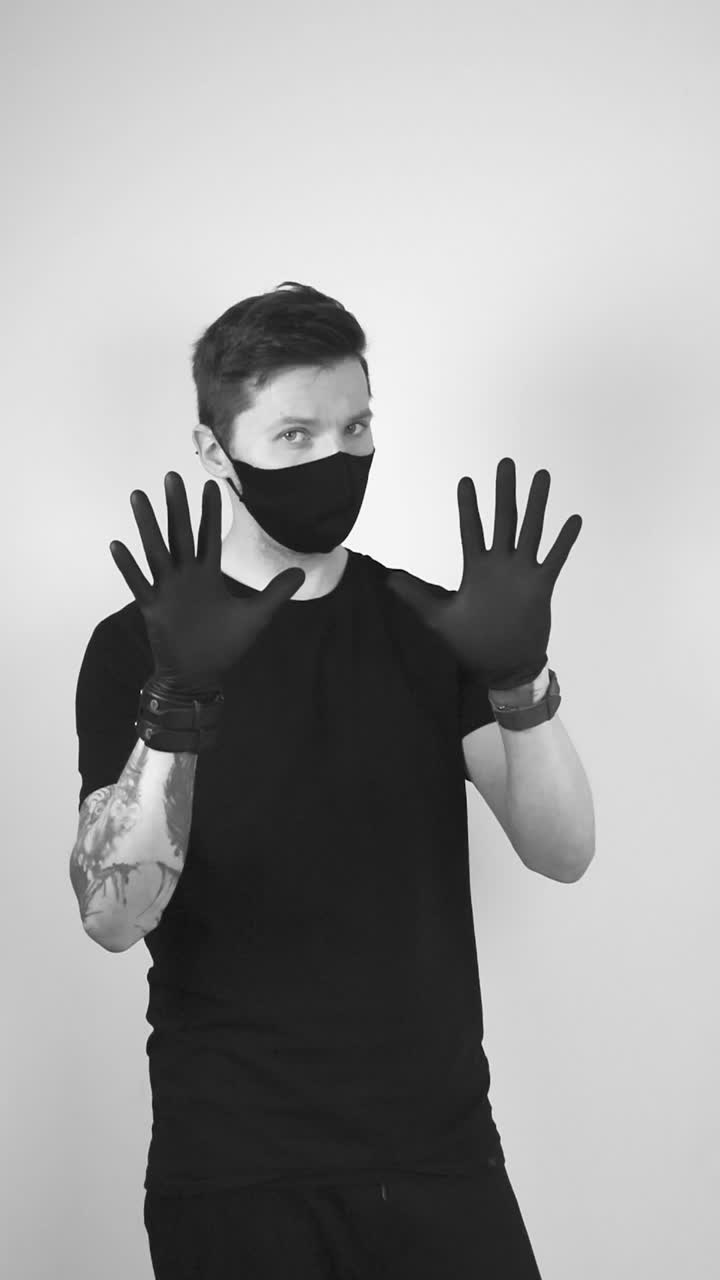 Grayscale footage of a tattooed man with medical protective black rubber gloves pointing to his covid face protective medical mask and showing also his gloves. Showing how and what to wear at pandemic