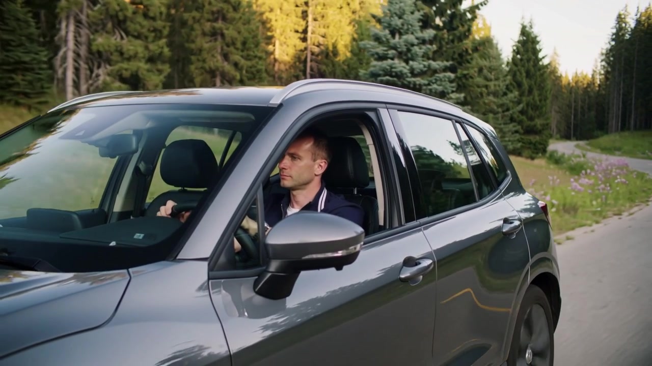 Uomo in polo blu navy che scruta la strada mentre si avvicina a una curva, alla guida di un SUV grigio su una strada forestale.
