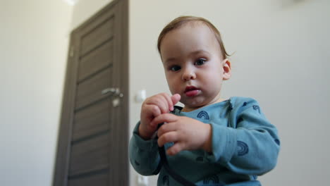 baby boy in blue shirt sits on the floor looking curiously on the shoe lace in his hands. cute toddler plays with a cord waving it cheerfully. low angle view close up.