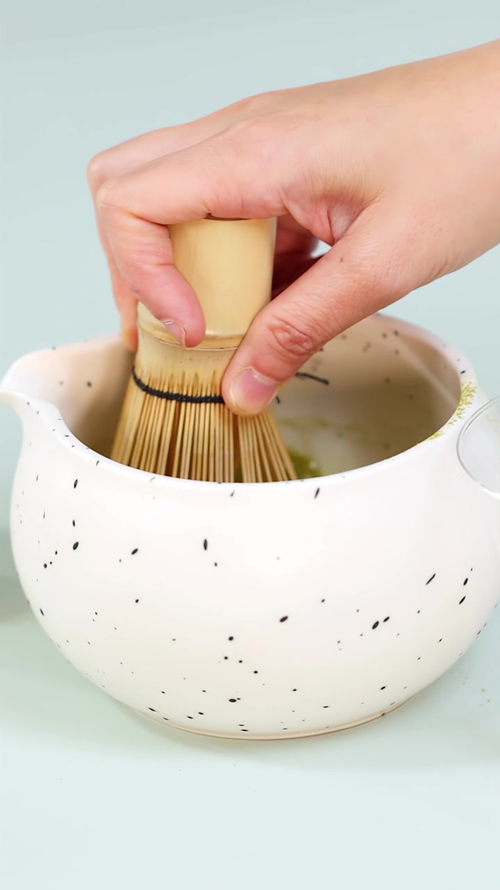 Primo piano di una mano che sbatte il tè verde matcha in una ciotola di ceramica maculata sotto una luce soffusa
