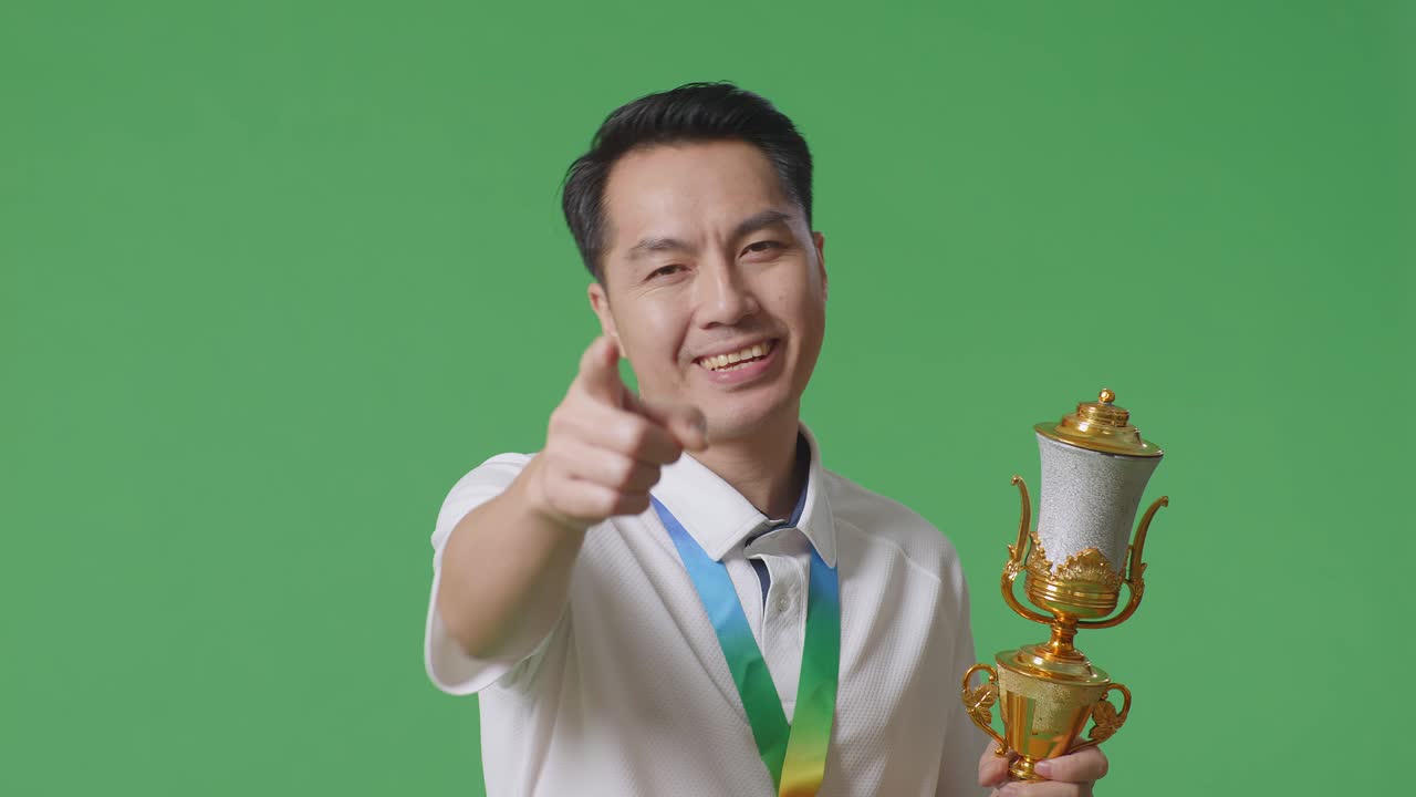 금메달과 트로피를 들고 있는 아시아인 남자의 가까운 사진, 지표손가락 표시 없이 불쾌감을 표하고 스튜디오의 녹색 스크린 배경에서 카메라를 향해 미소 짓고 있다.