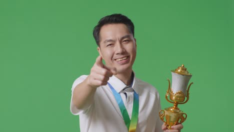 close up of asian man with a gold medal and trophy disapproving with no index finger sign and smiling to camera on green screen background in the studio