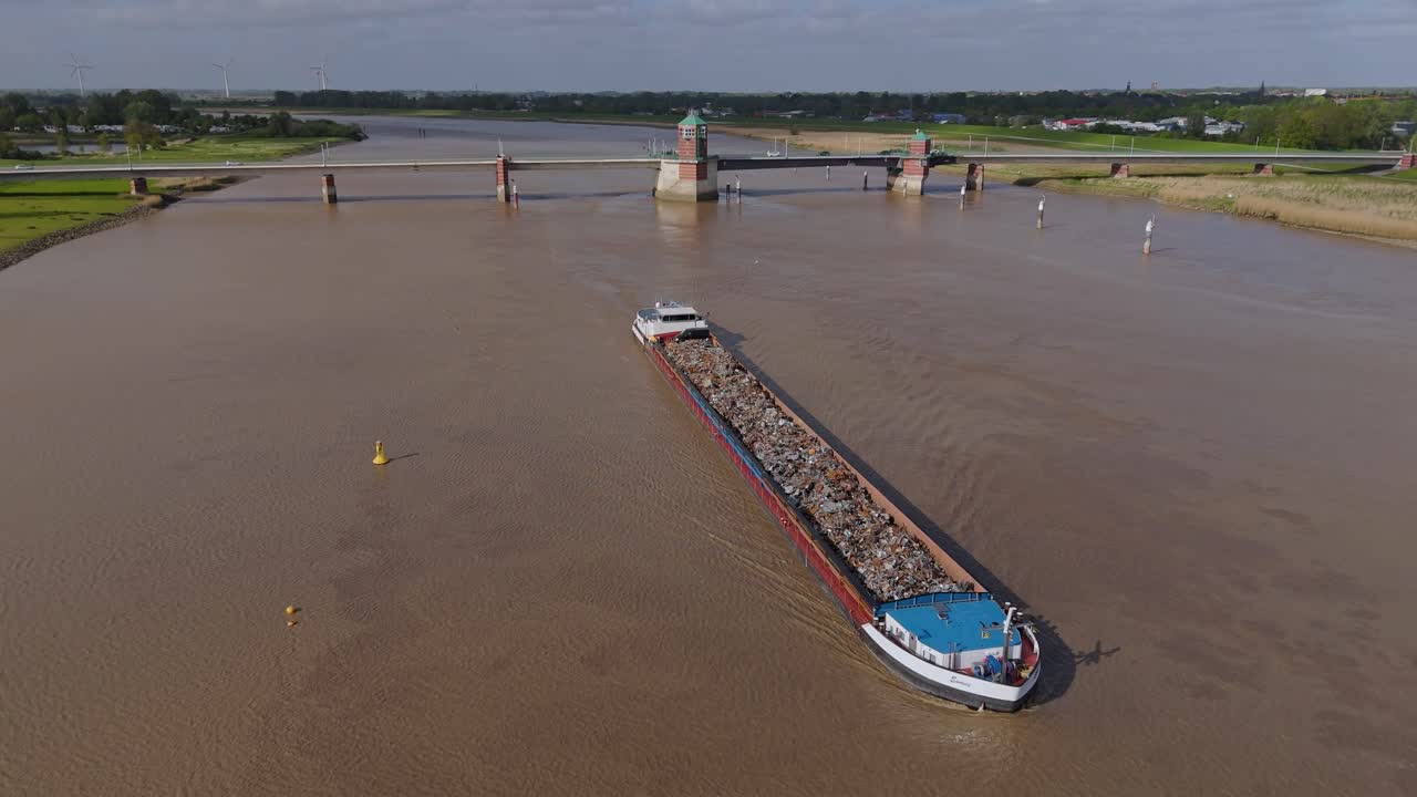 A cargo vessel travels under the Jann-Berghaus Bridge on the Ems River in northern Germany, captured by drone. Scenic view with traffic, green dike landscape and shipping route in East Frisia.