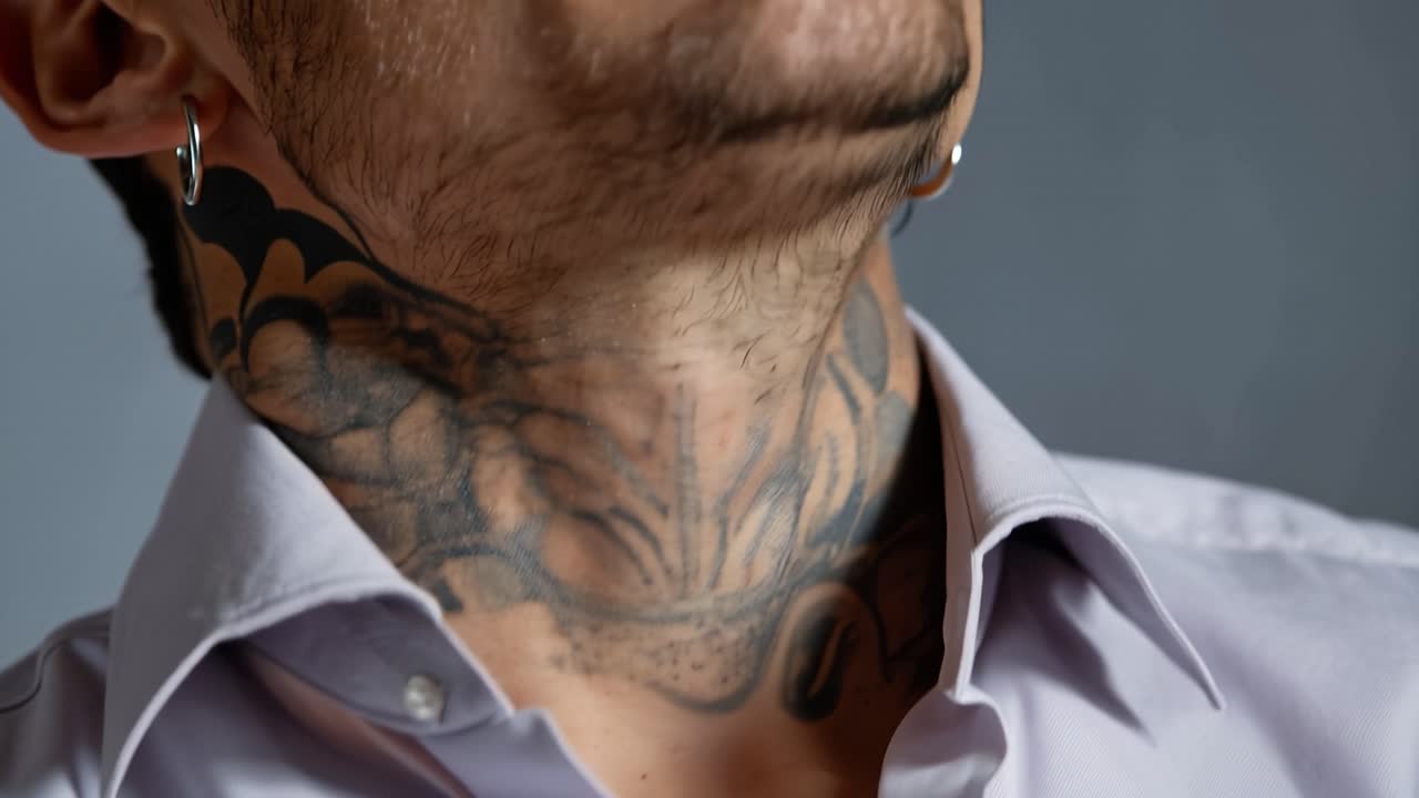 Close-up of a man's neck and chin. Tattooed skin shows intricate, dark patterns. Light highlights his facial hair and ear ring. Expression is calm and composed. Ideal for fashion, tattoo
