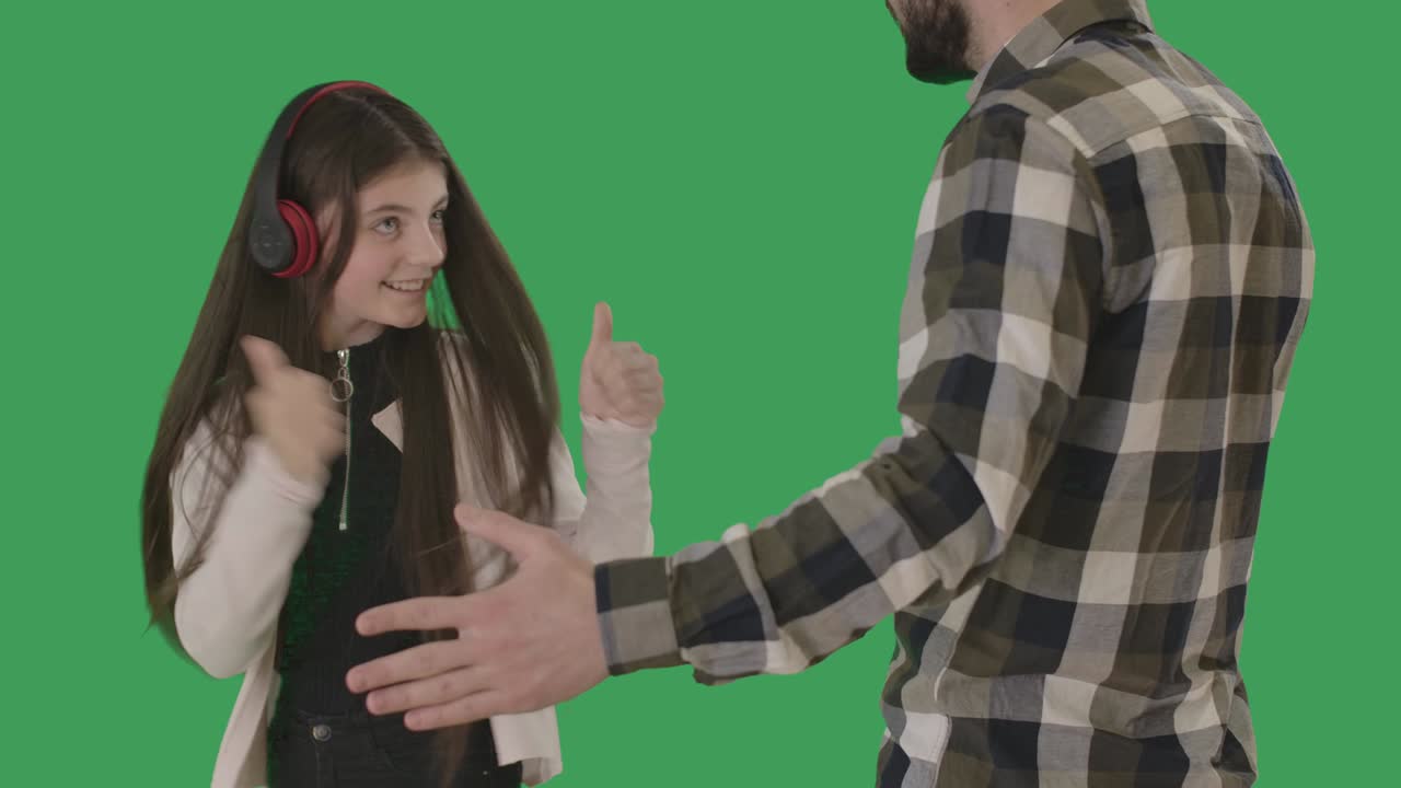una adolescente caucásica traviesa dirigiéndose a su padre regañándola y mostrando los pulgares hacia arriba. una chica alegre con auriculares ignorando el conflicto con un hombre nervioso. chromakey, pantalla verde. cine 4k prores hq.