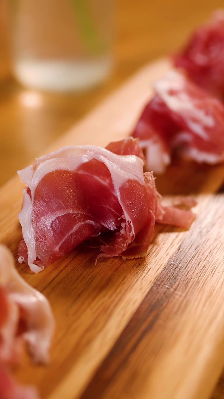 Ripresa ravvicinata in movimento di fette di prosciutto pregiato disposte su un tagliere rustico in legno.
