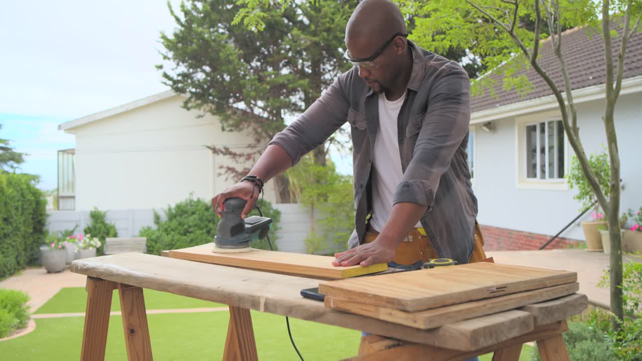 Homme afro-américain mettant en marche une ponceuse, stabilisant une planche et la ponçant, préparant ainsi la planche dans le jardin.