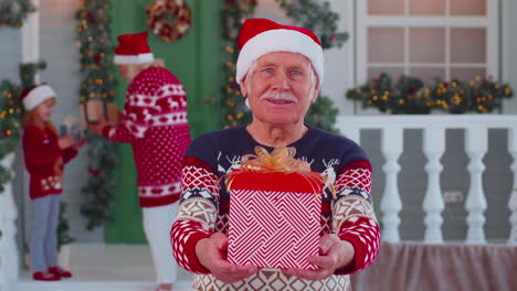 Portrait-of-grandfather-man-presenting-gift-box-smiling-near-decorated-Christmas-house-with-family