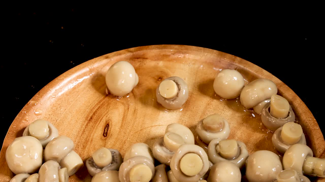 Whole button mushrooms fall onto a round wooden plate against a black background, captured in high-speed close-up with dramatic lighting and minimal camera movement