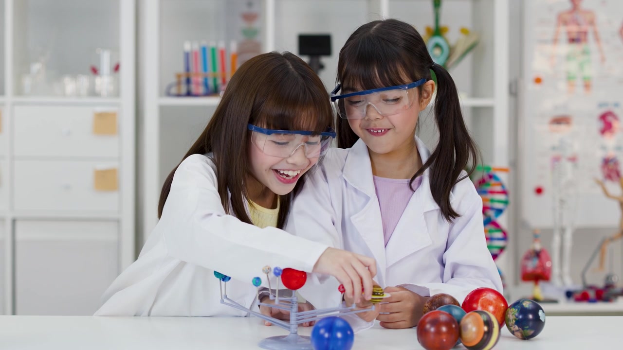 Kaksi nuorta tyttöä suojalasit ja laboratoriotakit päällään tutkii planeettamallia koulussa.
