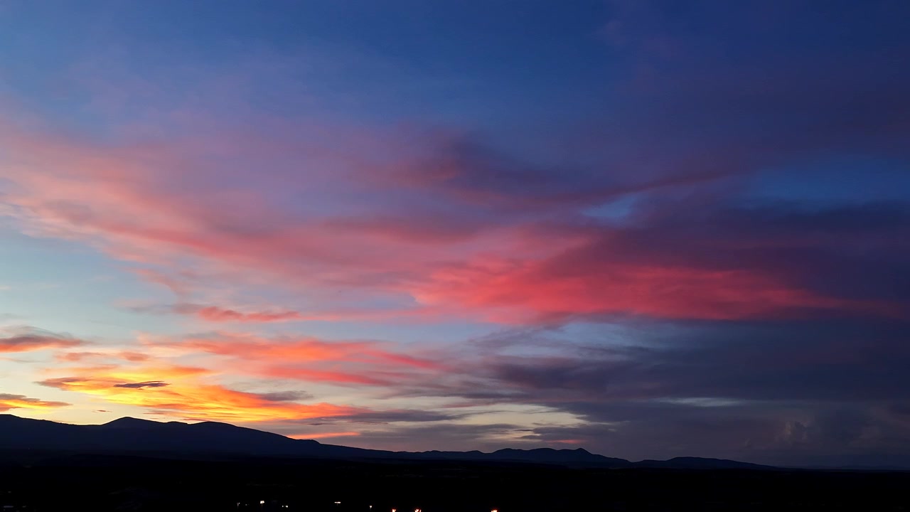 4K drone hyperlapse of vibrant, rainbow-colored clouds fading as dusk transitions to night. Town lights begin to flicker below a dark, silhouetted mountain ridge under the shifting evening sky.