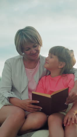 mujer de mediana edad y niña de top rosa leen juntos en una pala de heno, la mujer abraza a la niña estrechamente con un sombrero de paja cerca, en un vasto campo abierto