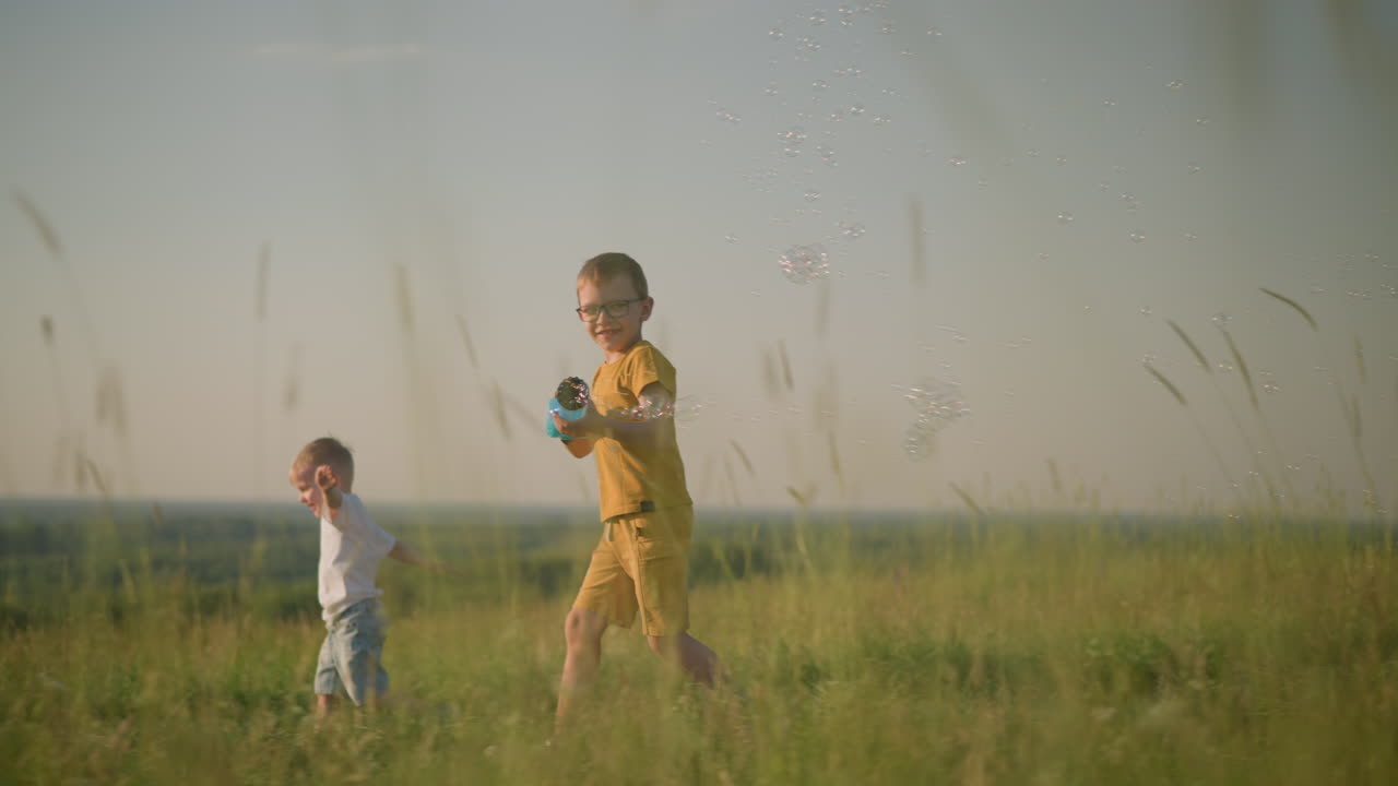 A young boy wearing a yellow shirt and shorts, with glasses, runs through a grassy field while holding a plastic bubble gun that releases bubbles. younger brother,in white, runs beside him laughing