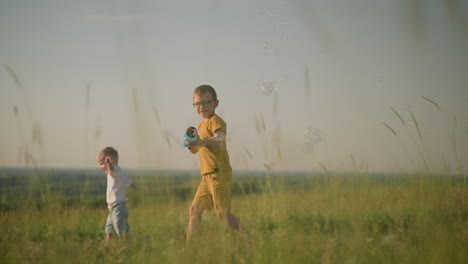 a young boy wearing a yellow shirt and shorts, with glasses, runs through a grassy field while holding a plastic bubble gun that releases bubbles. younger brother,in white, runs beside him laughing
