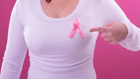 Video-of-midsection-of-caucasian-woman-wearing-pink-cancer-awareness-ribbon,-with-pink-background