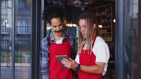 two diverse male baristas wearing aprons standing in the doorway and using digital tablet