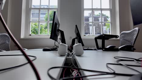 a contemporary office environment featuring multiple computer setups on desks, with large windows providing natural light and a view of an outdoor landscape