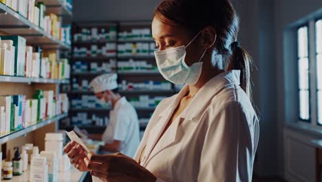 female pharmacist wearing surgical mask carefully reading prescription, male colleague working in background at modern drugstore, showcasing healthcare professional environment during pandemic